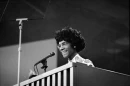 A black and white photo of Shirley Chisholm. She is standing behind a podium with a microphone and is smiling.