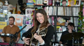  A woman smiles brightly while playing an acoustic guitar and standing behind an office desk, with two musicians out of focus in the background.