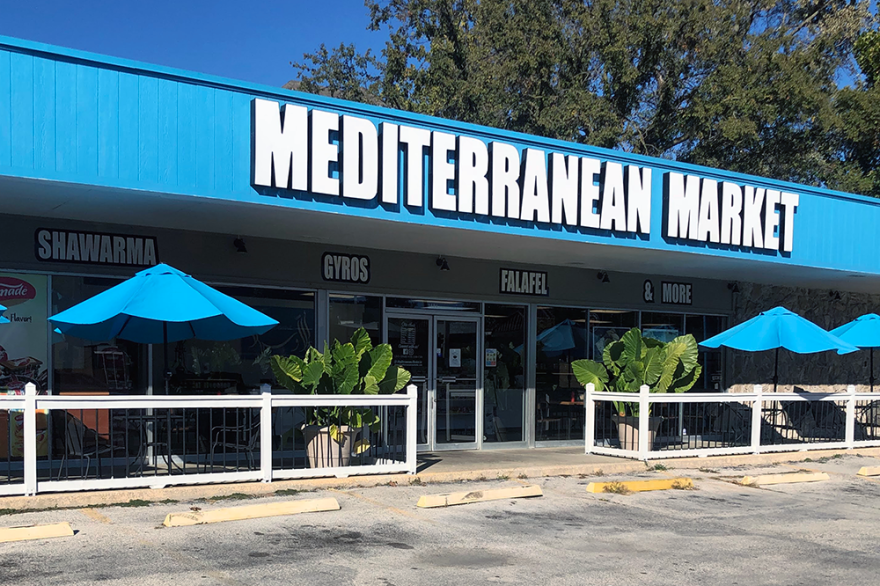 Exterior view of Mediterranean Market, with a blue awning and blue umbrellas and potted palm plants in front.