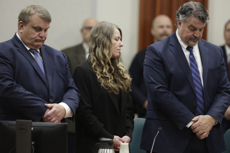 A woman with long blonde hair, wearing all black, standing between two men. All three of them have their hands crossed in front. 