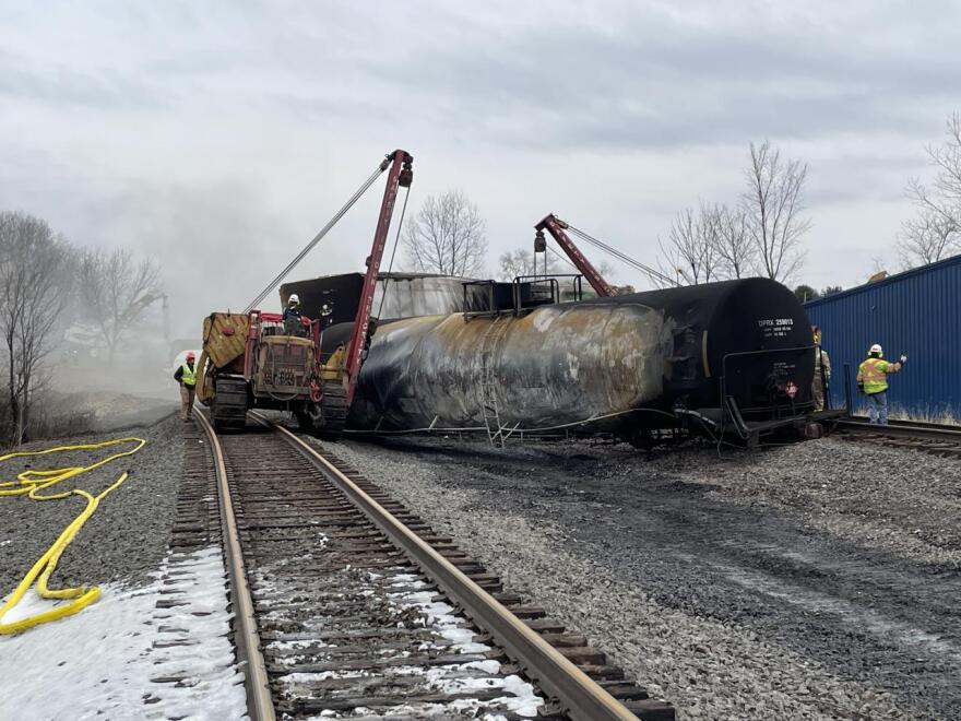 Two small cranes lift a corroded tanker car from between two sets of railroad tracks. Workers in helmets and high-visibility vests walk beside the wreck.