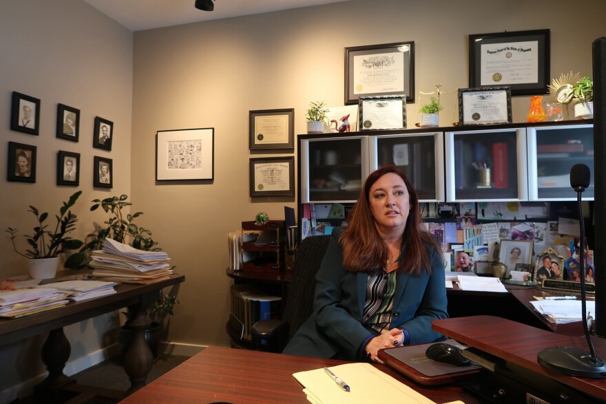 Elisabeth Trefonas sitting at her desk. Pictures and certificates hang on the wall. Behind her, photos and cards are tacked to the wall in a big collage.