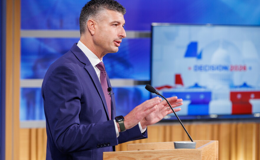 A man in a suit speaks at a podium in front of a TV that reads "Decision 2026"