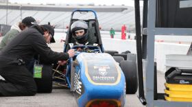 Students kneel next to a blue racing car with a person in the car.