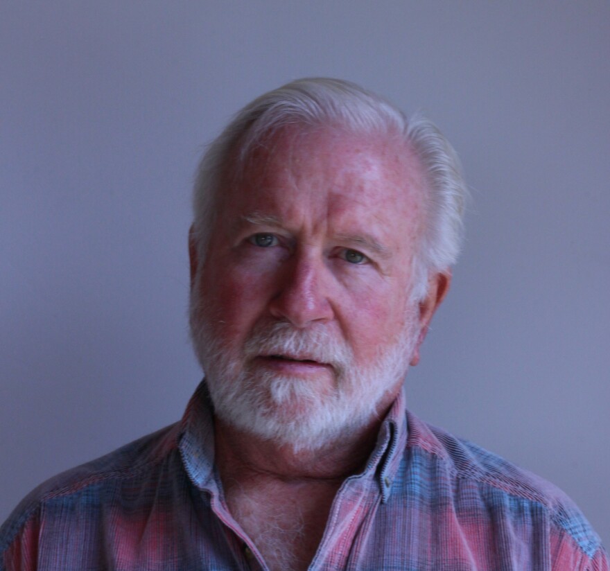 Brent Cook at this StoryCorps appointment in Springdale, Utah.  He gazes at the camera with rather a bemused expression.  He has short white hair parted on his left, and a trim, white beard and mustache.  He wears a red and blue plaid button-up shirt, with two buttons open.