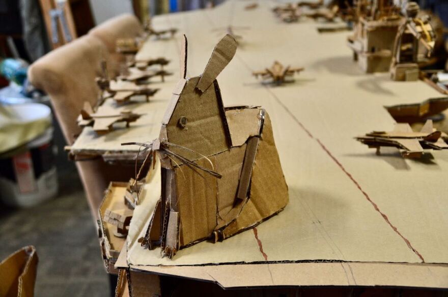 A cardboard rabit sits on a scultpure of a cardboard ship.