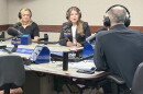 Three people wearing headphones sit at a table in a radio talk studio: a woman at left has long blonde hair and is wearing a short-sleeved black shirt and beaded necklace; a woman at center has long brown hair and is wearing a navy blazer over a white top; a man with very short dark hair at right is sitting with his back to the camera and wearing a navy shirt.