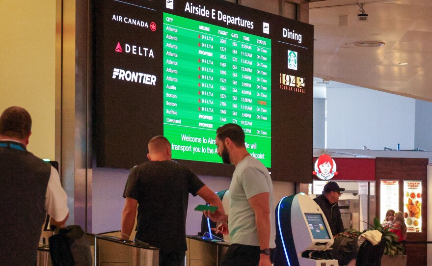 Travelers pass through an airport security gate while a giant screen behind them shows flight statuses. Only one of them appears to be delayed.