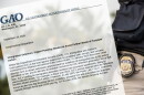 First page of the GAO report sits on top of a photo with an ICE agent's badge and gun in front of blurry palm trees. 