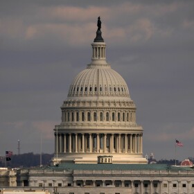 The United States Capitol building in Washington, D.C., with its dome and the Statue of Freedom perched on top. The sky in the background is filled with dark clouds.