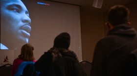 Three people sit in the foreground silhouetted against a large projection screen where the closeup face of Dr. Martin Luther King, Jr. is projected.