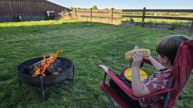 A young boy wearing a Hawaiian-print shirt sits in a red camp chair in front of a small fire in a metal fire pit. He's about to bite into a hot dog he just cooked over the flames.