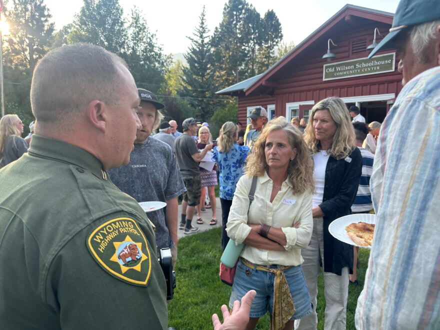A uniformed Wyoming Highway Patrol Officer speaks with his hands to a small circle of people, who don't look pleased. They're among a large crowd standing outside a brown building.