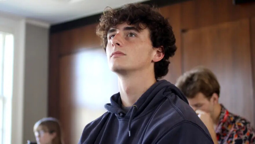 Young people in a room with wood paneling. A young man in a dark blue hoodie is in the foreground looking up.