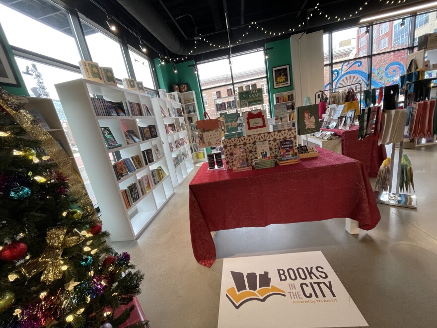 A display of books sits atop a table with a red tablecloth. Behind it, white shelves lined with books. On the left of the frame, a decorated Christmas tree. On the floor, a sign reads "Books in the City."