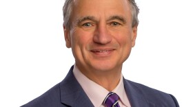 A man with short grey hair is wearing a navy blazer, pink button-down shirt and purple striped tie.