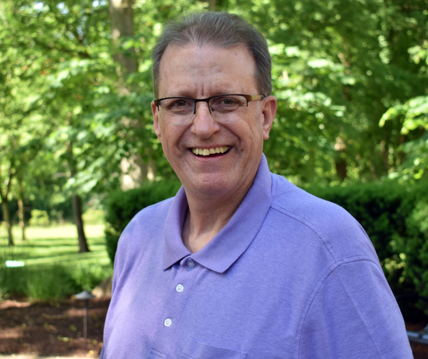  A man wearing glasses smiles at the camera while standing outdoors in front of trees.
