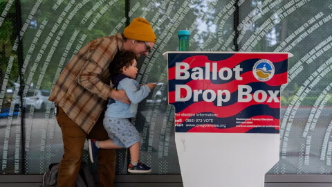 FILE - Matt lifts up his three-year-old son, Theo, to cast his ballot into the Hillsboro Public Library-Shute Park ballot drop box in May 2026. Election officials are increasingly recommending voters plan to use drop boxes rather than mailing their ballot.