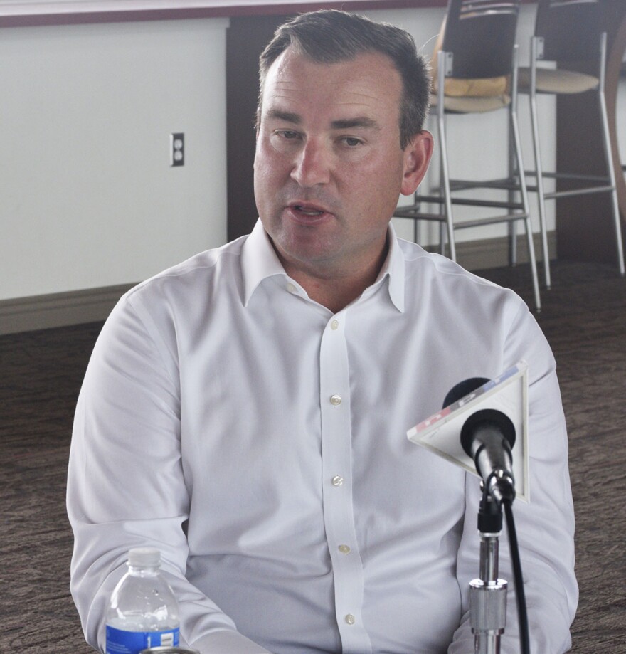 Man in a white buttoned-down collared shirt seated in front of a microphone and two stools placed against a wall behind him.