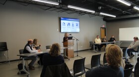 A group watches a blonde woman present at a podium at the front of a meeting room