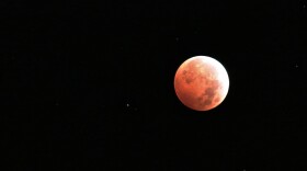 A red moon against a black background with a few white stars. 