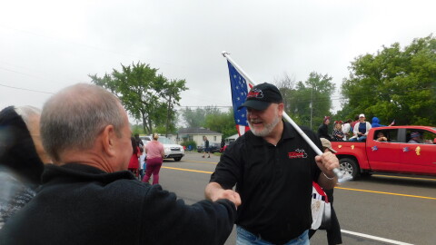 A white man wearing a black polo shirt and baseball cap and carrying an American flag shakes the hand of another white man along a parade route.