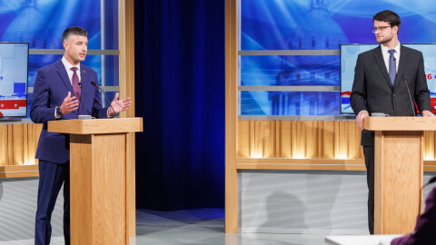 Two men in suits stand at podiums in a TV studio