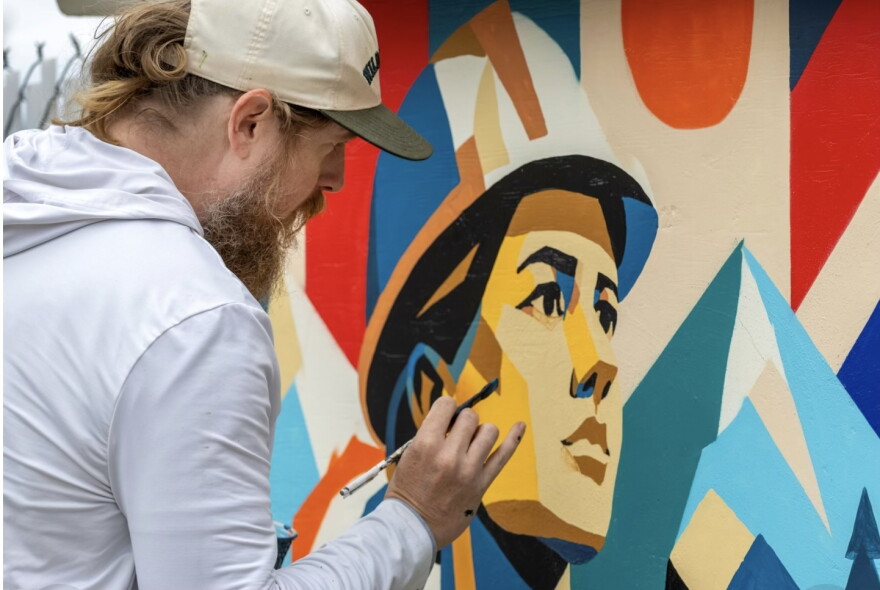 A close shot of the artist, a bearded white man, leaning towards his mural, painting the details of a face looking in the distance, and wearing a helmet. 