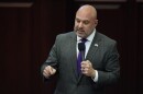 A bald man with a grey trimmed beard wearing a suit and tie, speaking into a microphone. 