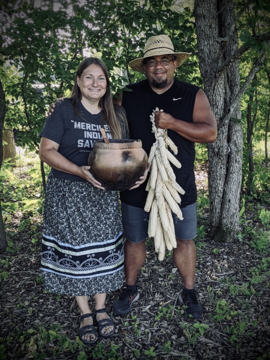 A man and woman stand together smiling at the camera. The man wears a straw hat and holds a braid of white corn. The woman holds a clay cooking pot. 