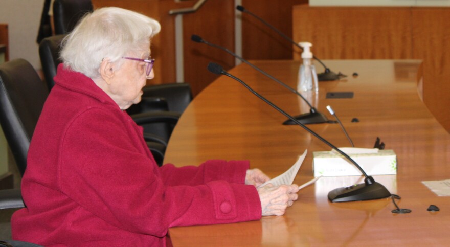 An older woman with white hair, wearing purple glasses, and a bright red coat reads from a speech on paper. She's sitting in front of a microphone set on a large wooden desk. 