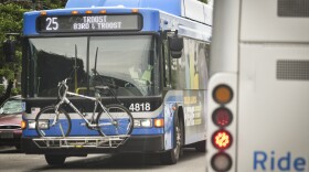 The front of a blue bus travels toward the camera on a city street.  On the front, the route marquis reads "25 Troost 83rd and Troost."