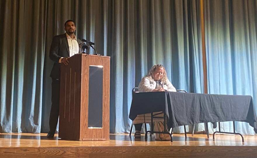 Abraham Morris stands behind a podium on stage as he gives a presentation at South Division High School. The moderator is sitting behind a table next to him.