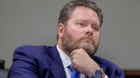 Man in a blue suit, brown hair and beard sitting down and looking to the right