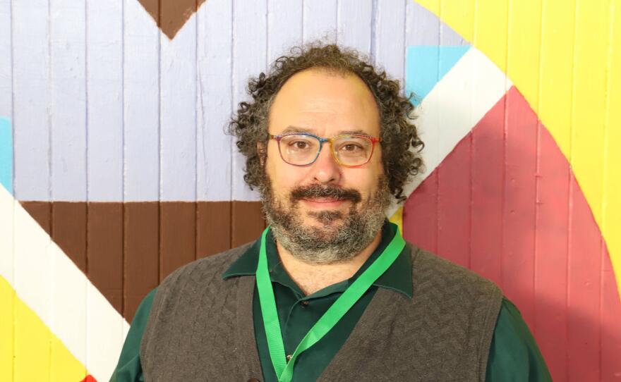 A man with curly brown hair and a brown beard is wearing glasses, a green button-down shirt, brown sweater vest and green lanyard while standing in front of a multi-colored wall.