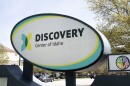 The outside oval sign of the Discovery Center of Idaho near its parking lot. It is white, with black lettering and yellow/turquoise highlights. 