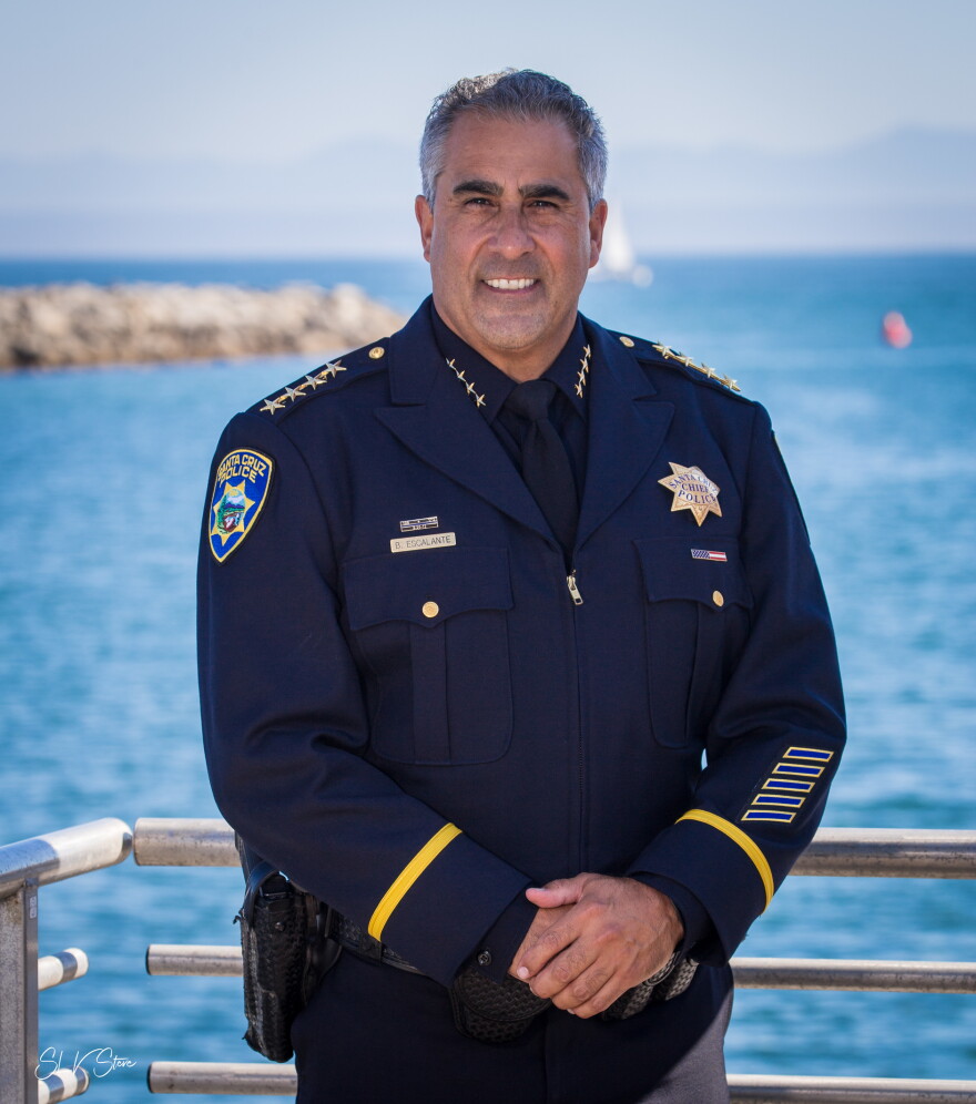 A man in a formal blue police uniform stands with his hands clasped in front of him. He's in front of a railing and behind that is the ocean with a jetty in the distance.