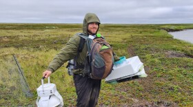 A person stands on a wetland carrying two large pieces of equipment and a backpack on his chest.