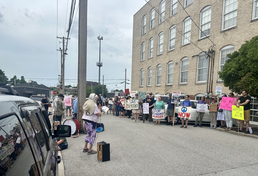 A few dozen protesters hold signs outside a tan brick building. A woman with purple leggings speaks to the small crowd. 