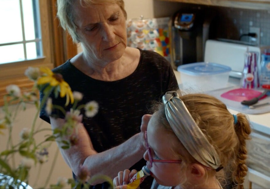 An older woman stands in her kitchen and fixes a young child's hairband.