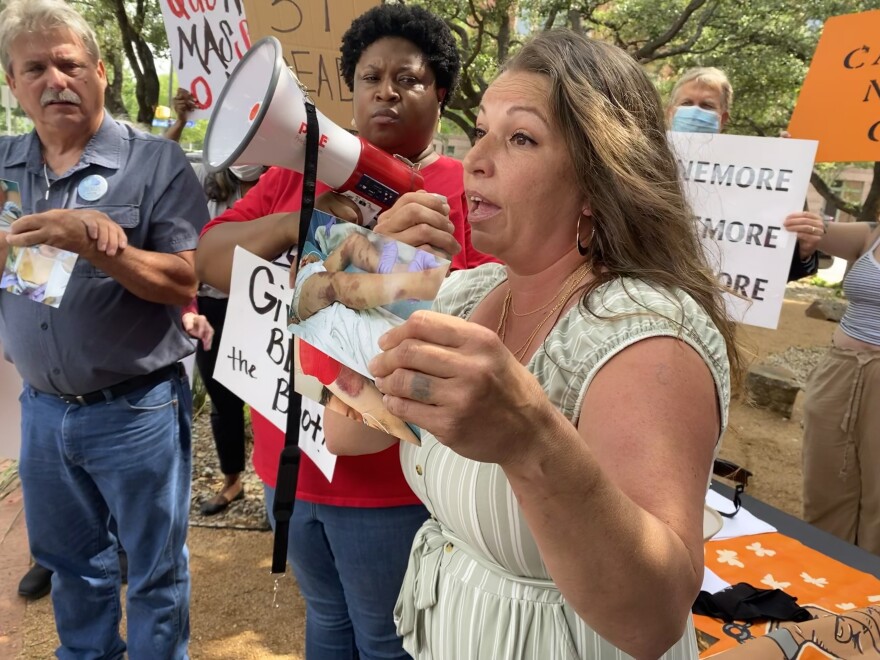 Kristina Salinas, a woman with long, light brown hair, holds up a photo of her sister's bruised legs. She stands before a group of people holding signs behind her at a protest.