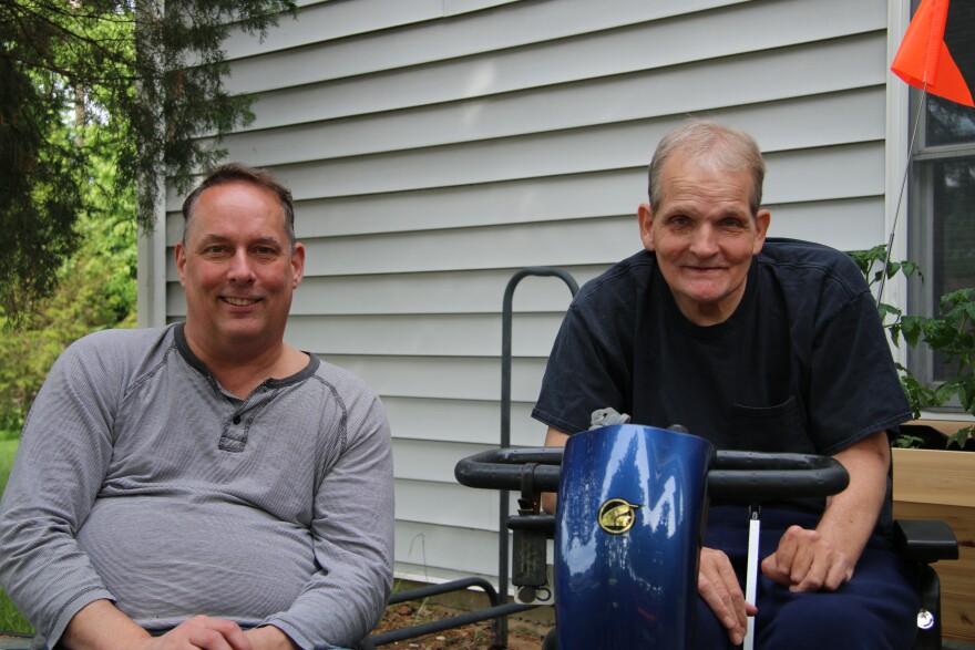 Chuck Graham, left, wears a gray shirt and smiles into the camera. His brother, Drew Graham, right, wears a blue shirt and sits in a bright blue power scooter. He also smiles into the camera. 