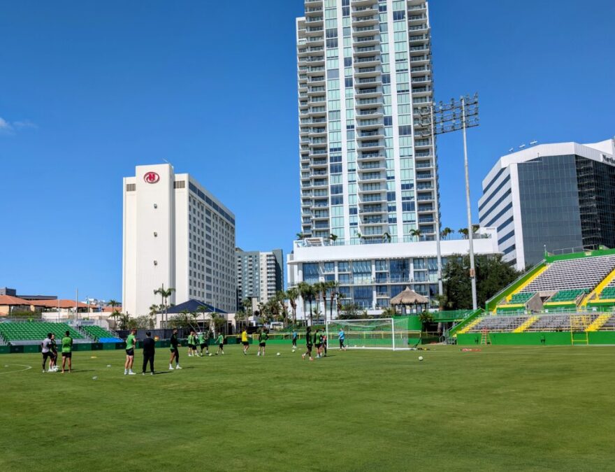 A wide shot of the Tampa Bay Rowdies field. Bleachers are to the right of the field. There is a Hilton hotel next to a large glass building. Players stand on the field.