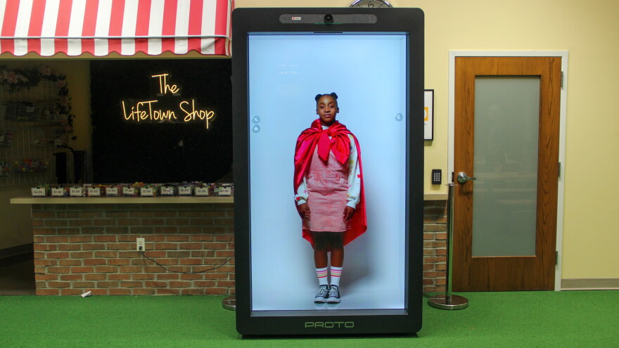 A lifesize hologram of a young Black girl wearing a cape stands in a large box labeled "Proto" next to a mock shop called The Lifetown Shop. 
