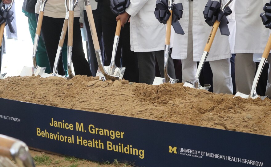 A line of shovels are standing in a pile of dirt, each decorated with a gift bow. The dirt has a sign in front of it for the Janice M. Granger Behavioral Health building.