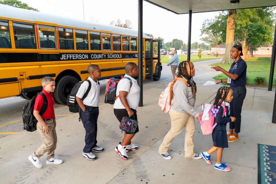 A woman with a lanyard speaks to a line of students entering a school building. A school bus is in the background.