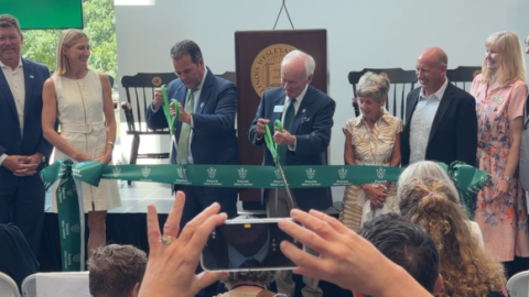 Two men in suits cut a green ribbon while several others look on
