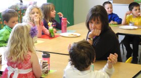 A woman sits in front of three wooden tables with seven younger children in front of her, eating lunch.