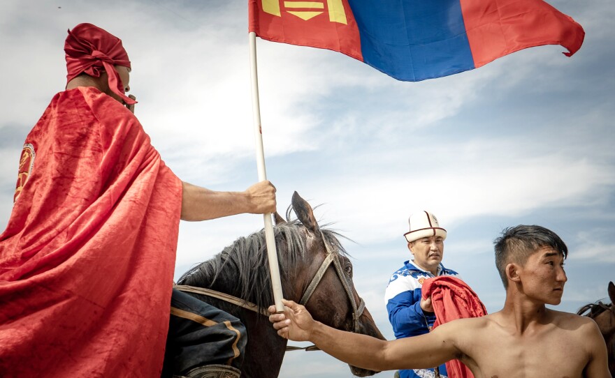 A member of the Mongolian horse wrestling team passes a flag to another member who is preparing for a victory lap around the stadium after a match victory.