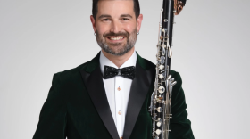 Un hombre de cabello oscuro muy corto, barba y bigote, vestido con un esmoquin negro, sostiene un clarinete bajo en su brazo izquierdo.